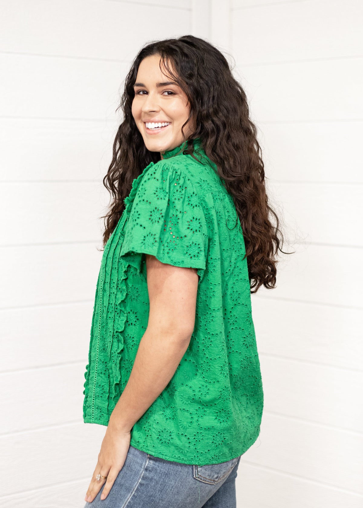 Woman with dark hair standing in a side profile position wearing the Lilly Top against a white background. The focal point of this image is the shoulder length and sneak peek of ruffles on the front of top. The model paired this top with dark-wash denim jeans. 