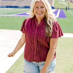 Woman with blonde hair standing in a front profile position wearing the Lilly Top in a lifestyle scene. The focal point of this image is the maroon eyelet pattern across top, ruffles along wooden buttons and high collar with ruffles. The model paired this top with light-wash denim shorts. 