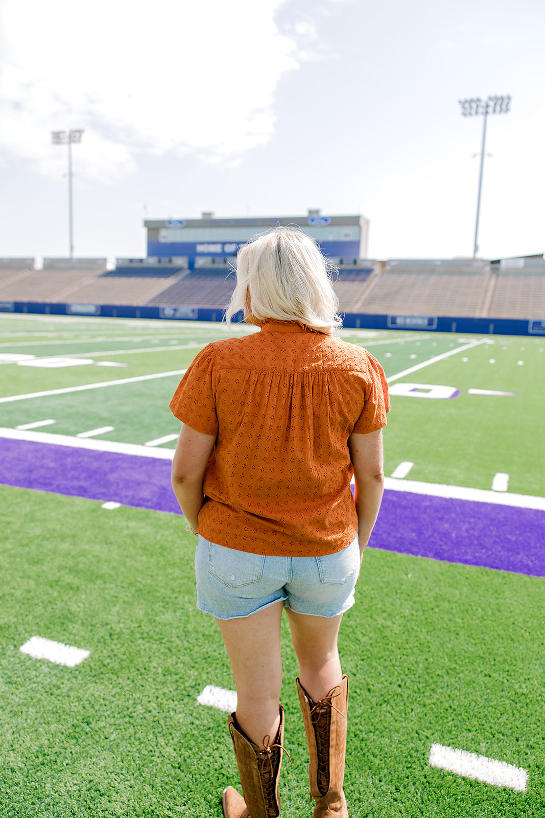 Woman with blonde hair standing in a back profile position wearing a short-sleeve top that's burnt orange eyelet pattern, and light-wash denim shorts. 