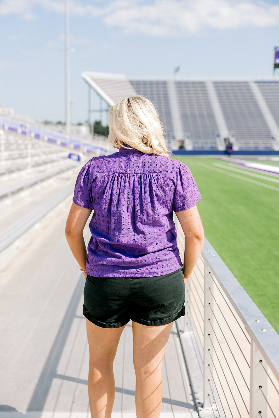 Woman with blonde hair standing in a back profile position wearing the Lilly Top in a lifestyle scene. The focal point of this image is the pleating on back of top and purple eyelet pattern. The model paired this top with black denim shorts. 
