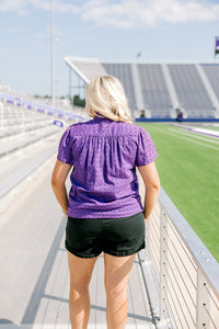 Woman with blonde hair standing in a back profile position wearing the Lilly Top in a lifestyle scene. The focal point of this image is the pleating on back of top and purple eyelet pattern. The model paired this top with black denim shorts. 