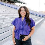 Woman with dark hair standing in a front profile position wearing the Lilly Top in the stands of a football stadium. The focal point of this image is the purple eyelet pattern, ruffles down side of buttons and collar. The model paired this top with black denim shorts.