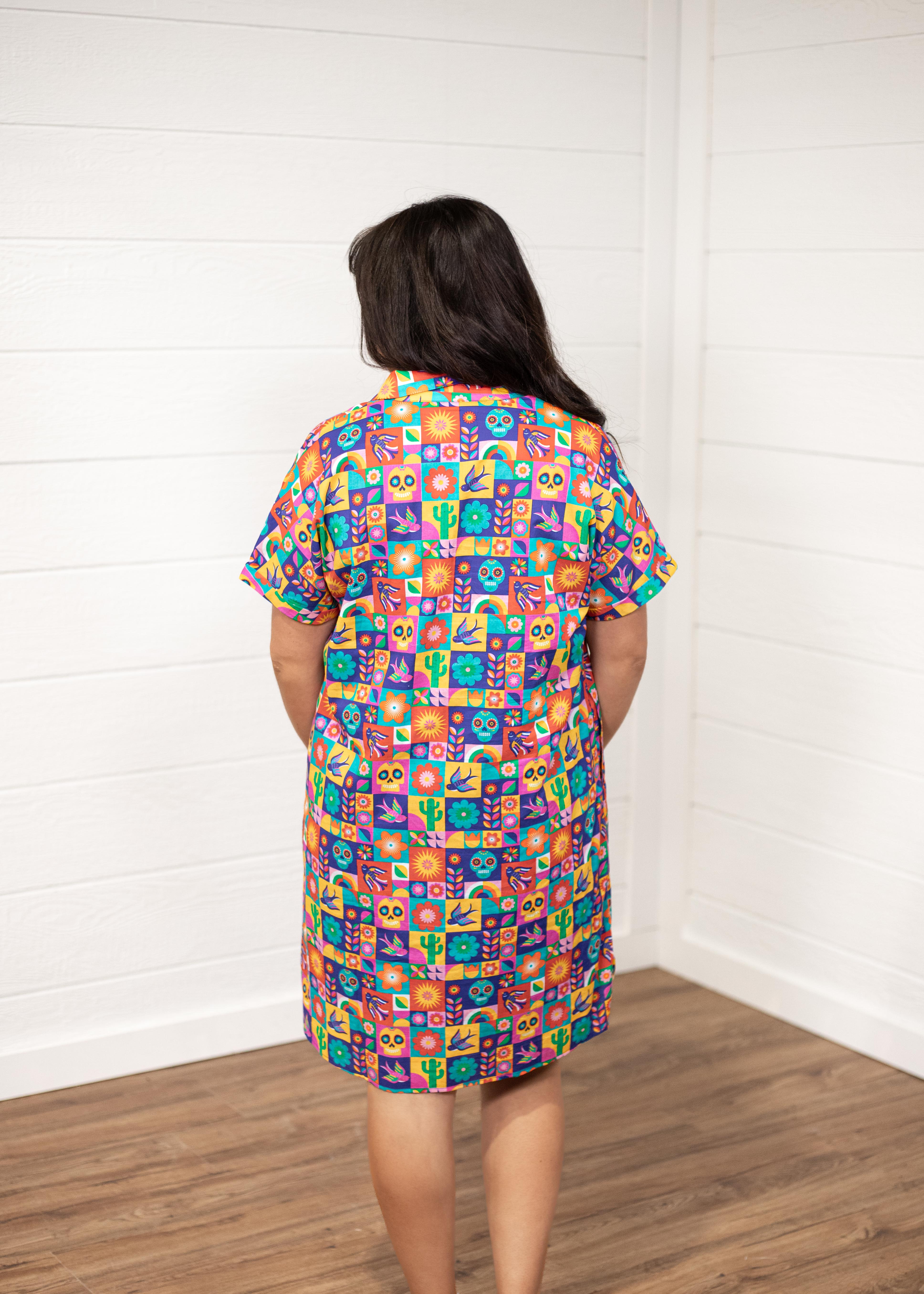 Woman with dark hair standing in a back profile position wearing the Lindsey Dress with a white background. The focal point of this image is the graphics on the back of the dress with slight emphasis on the collar and back of sleeves. 