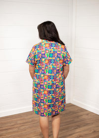 Woman with dark hair standing in a back profile position wearing the Lindsey Dress with a white background. The focal point of this image is the graphics on the back of the dress with slight emphasis on the collar and back of sleeves. 