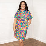 Woman with dark hair standing in a front profile position wearing the Lindsey Dress against a white background. The focal point of this image is the entirety of the dress with graphics visible. The model paired this dress with white and gold sneakers. 