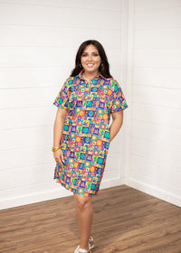 Woman with dark hair standing in a front profile position wearing the Lindsey Dress against a white background. The focal point of this image is the entirety of the dress with graphics visible. The model paired this dress with white and gold sneakers. 