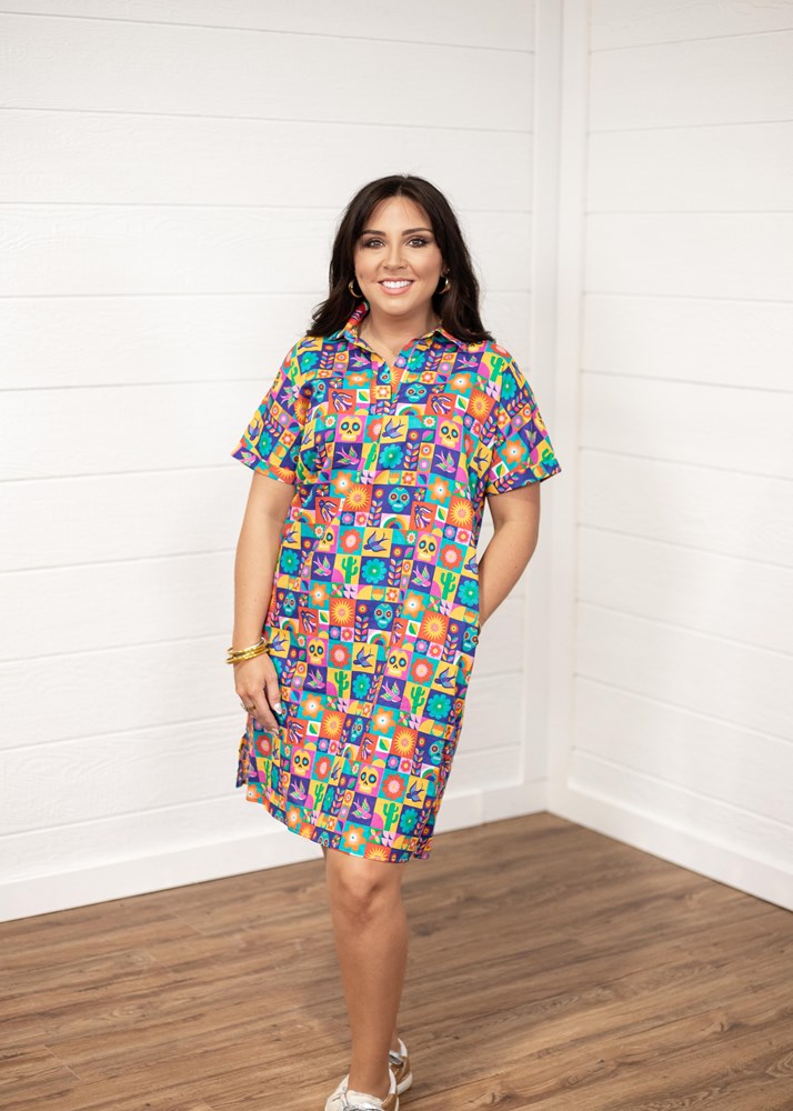 Woman with dark hair standing in a front profile position wearing the Lindsey Dress against a white background. The focal point of this image is the entirety of the dress with graphics visible. The model paired this dress with white and gold sneakers. 