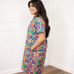 Woman with dark hair standing in a side profile position wearing the Lindsey Dress with a white background. The focal point of this image is the side graphics of the dress with emphasis on the sleeves. 
