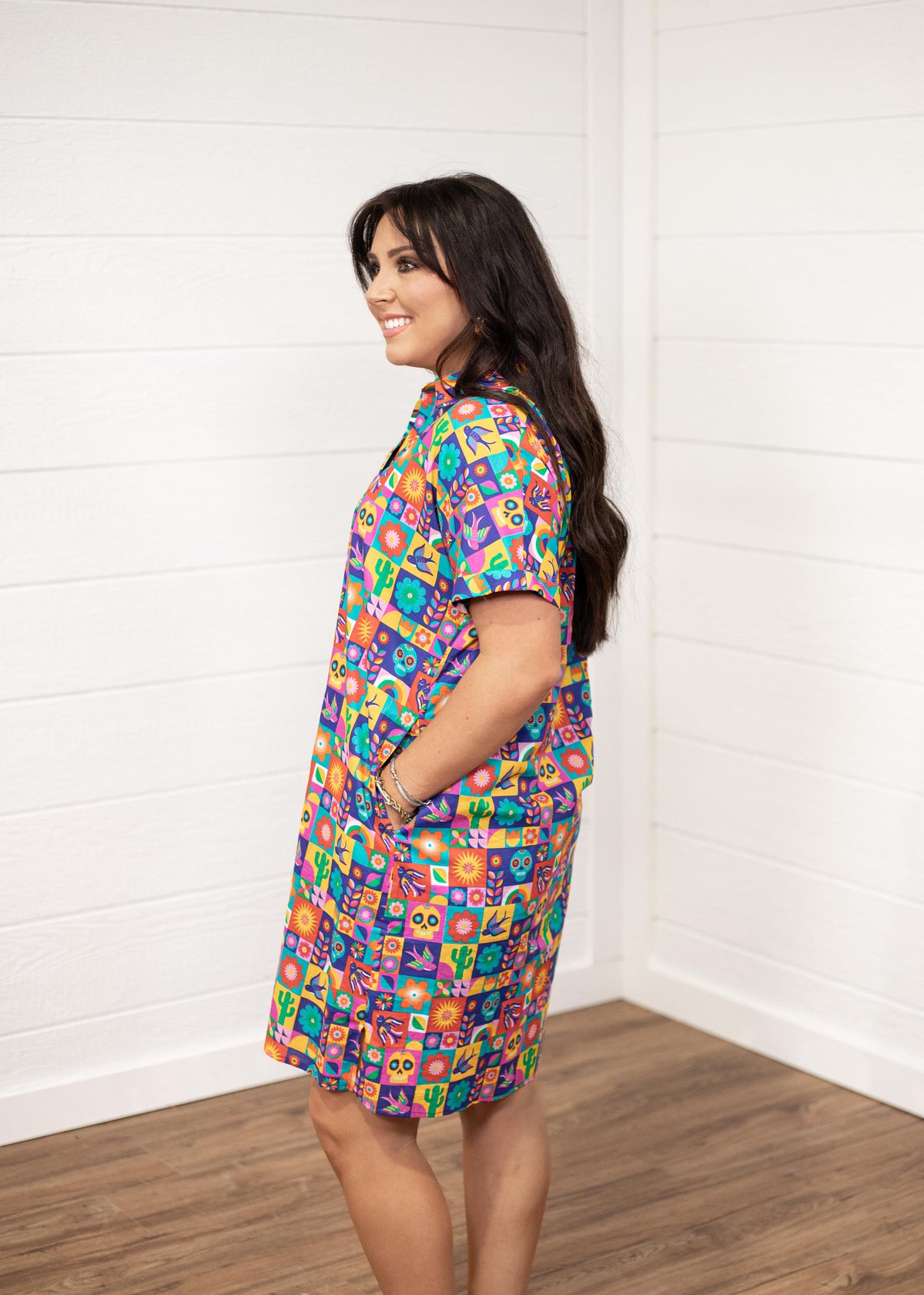 Woman with dark hair standing in a side profile position wearing the Lindsey Dress with a white background. The focal point of this image is the side graphics of the dress with emphasis on the sleeves. 
