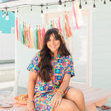 Woman with dark hair sitting in a front profile position wearing the Lindsey Dress with a backyard party styled background. The focal point of this image is the dress in bright/colorful scenery. 