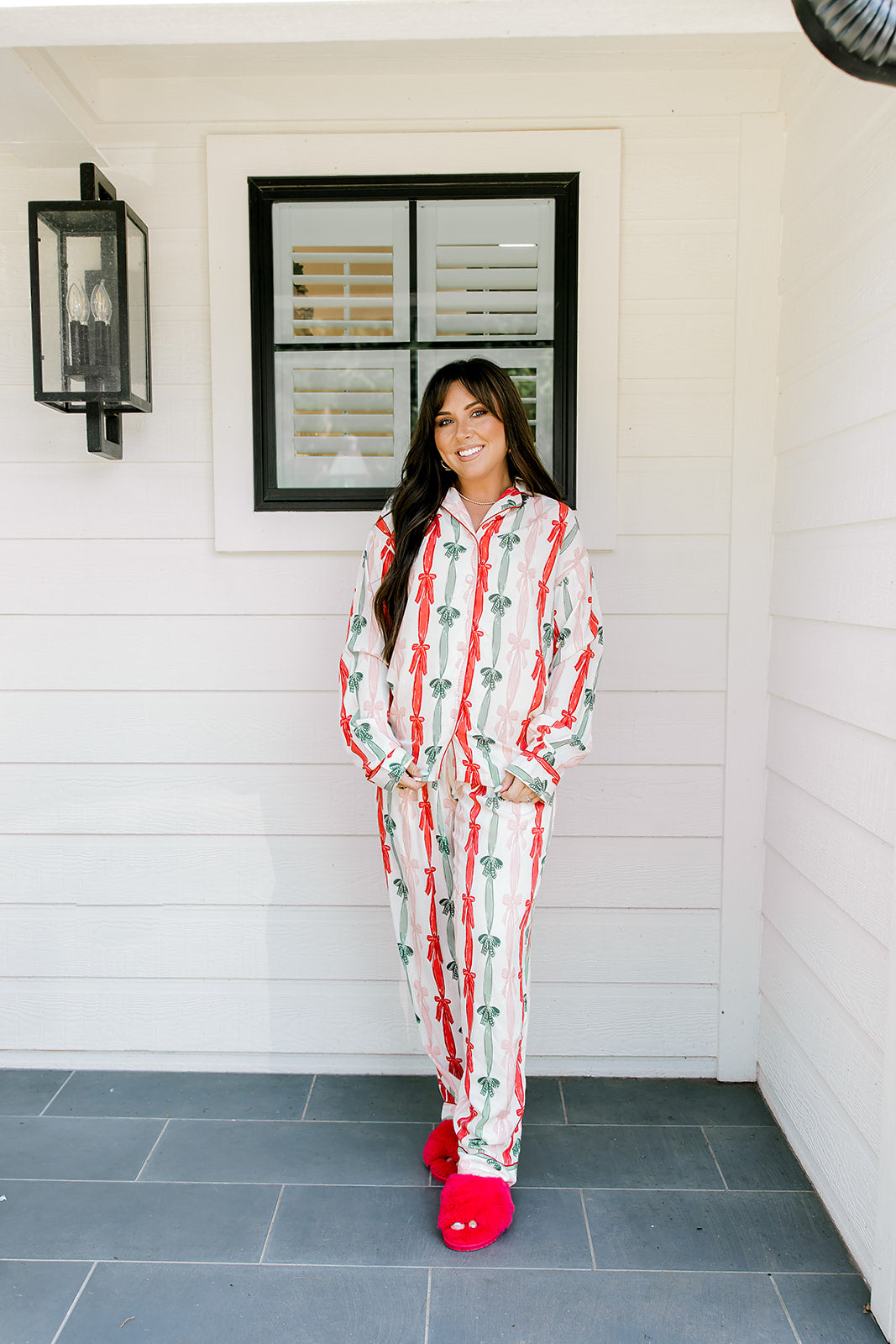 Woman with dark hair wearing a long-sleeve pajama top and pants set that features green, red, and pink bow graphics. 