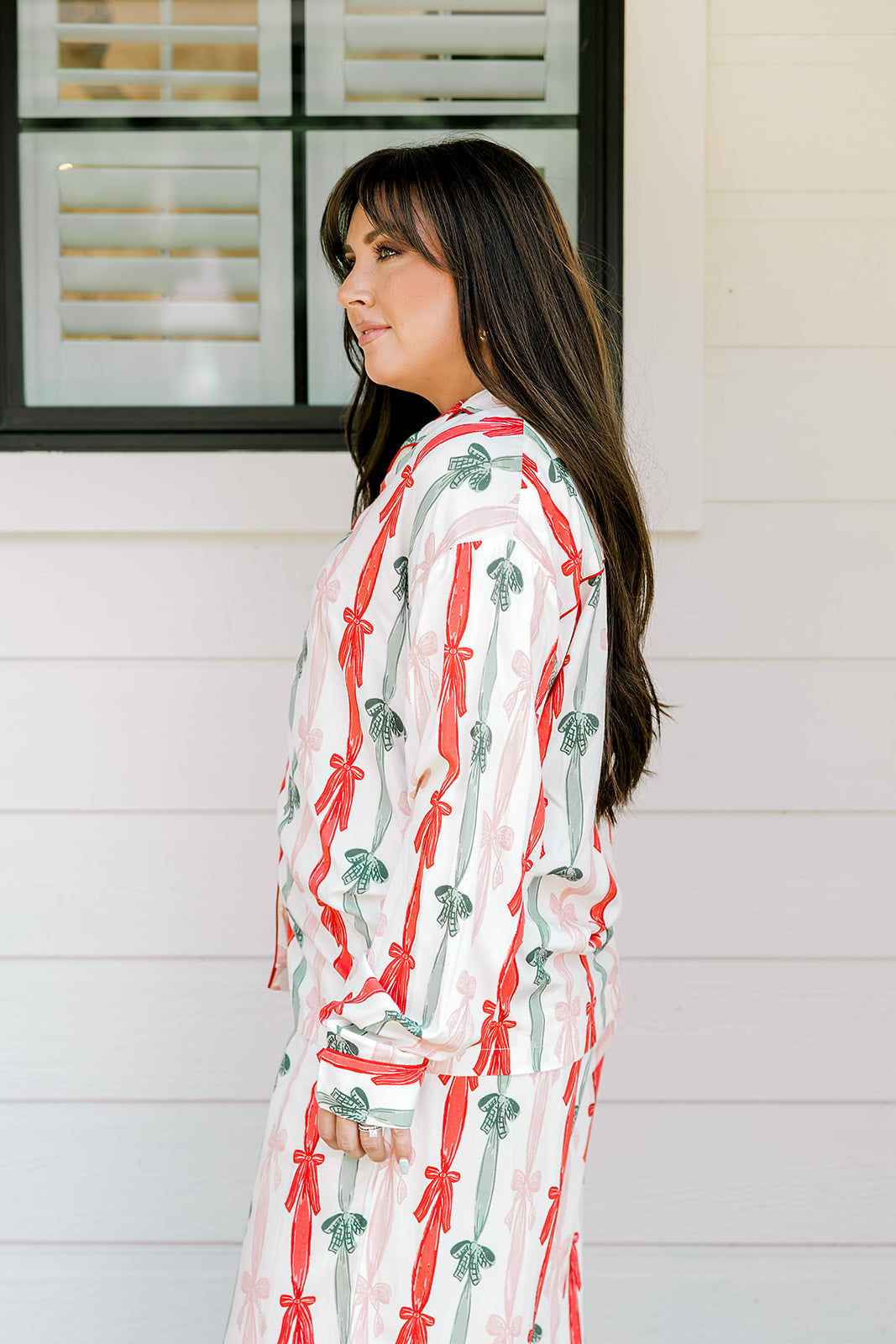 Woman with dark hair wearing a long-sleeve pajama top and pants set that features green, red, and pink bow graphics. 