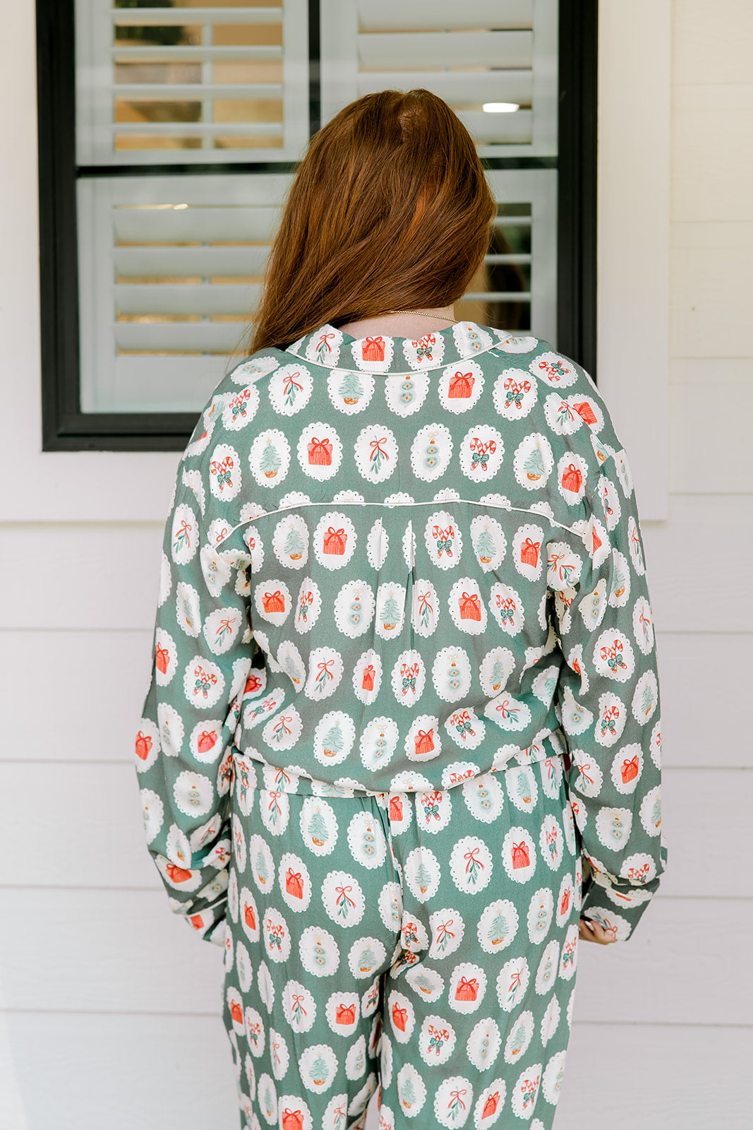 Woman with red hair wearing a long-sleeve top and long pant pajama set. The Pajamas feature fun red & green Christmas icon graphics. 