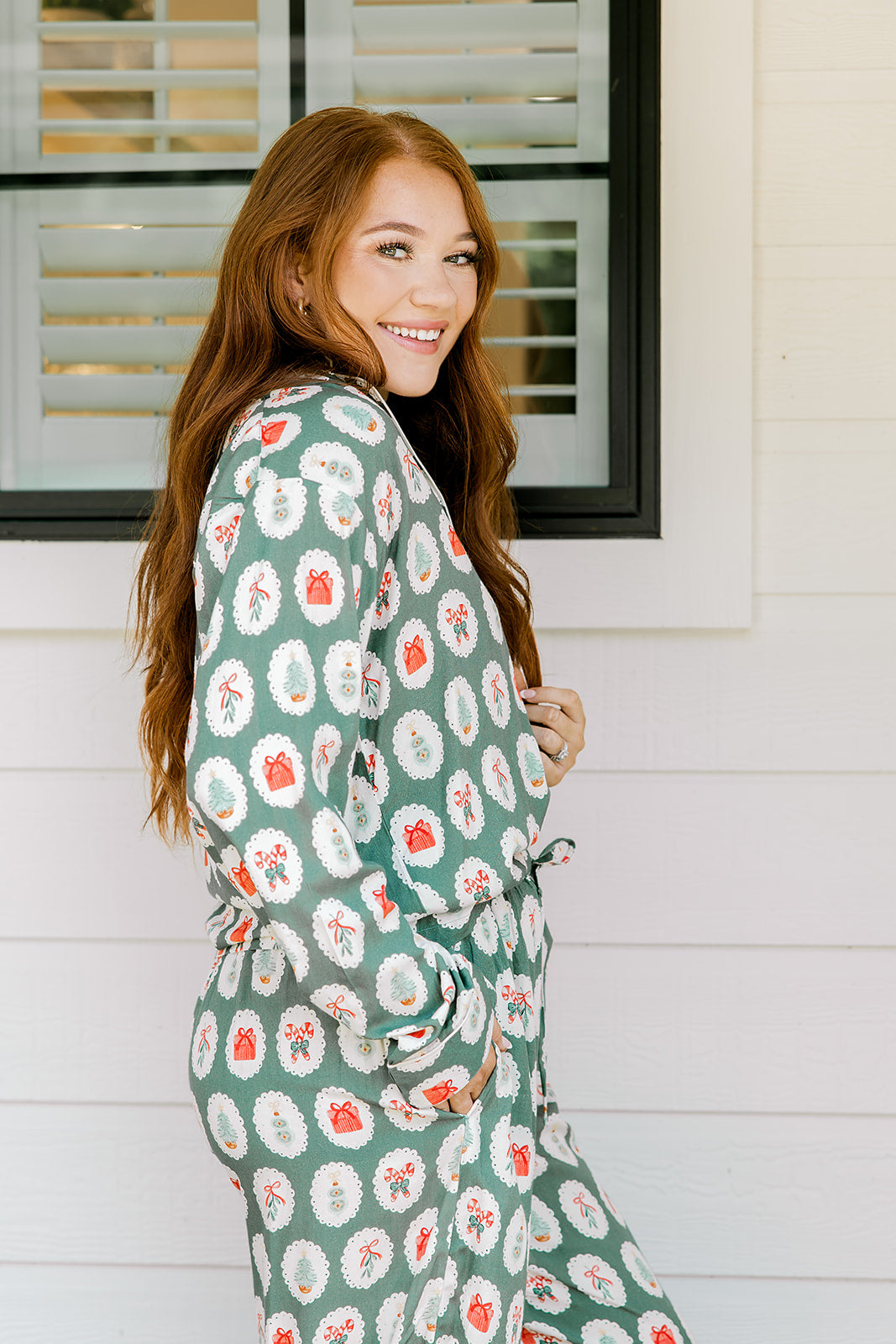 Woman with red hair wearing a long-sleeve top and long pant pajama set. The Pajamas feature fun red & green Christmas icon graphics. 