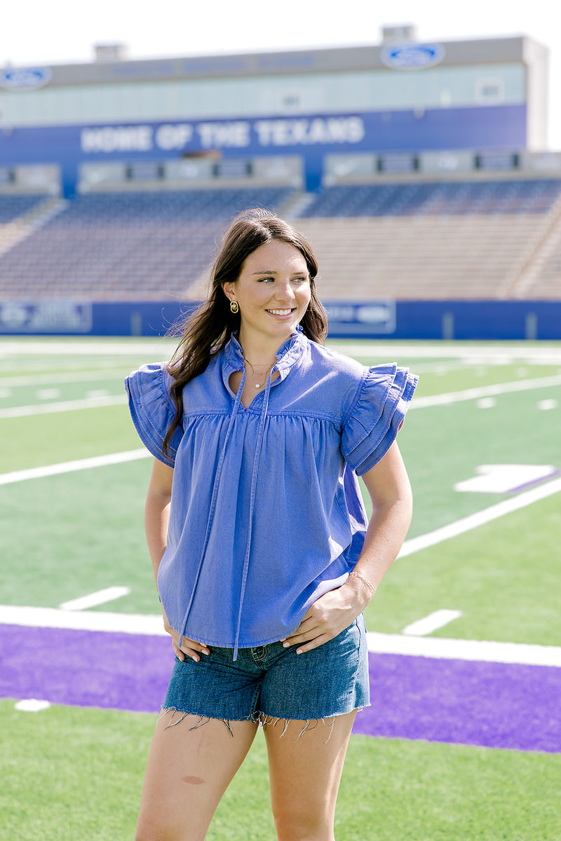 Woman with dark hair standing in a front profile position wearing the Marley Top in a lifestyle scene. The focal point of this image is the purple denim fabric, pleating on front of top, ruffles on sleeves, and tie in the front. The model paired this top with dark-wash denim shorts. 