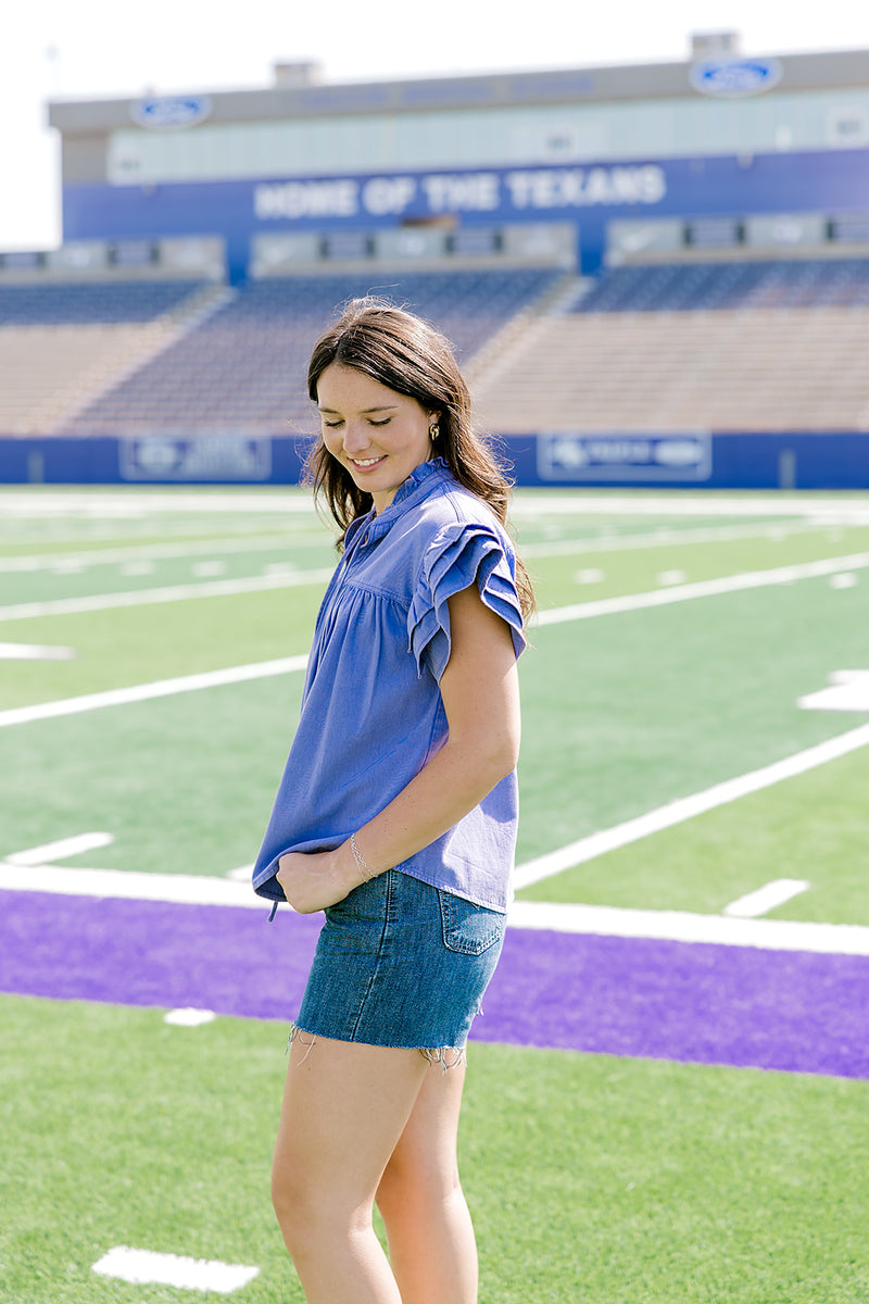 Woman with dark hair standing in a side profile position wearing the Marley Top in a lifestyle scene. The focal point of this image is the purple denim ruffled sleeve. The model paired this top with dark-wash denim shorts. 