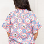 Woman with dark hair standing in a back profile position wearing the Mia Top against a white background. The focal point of this image is the intricate blue and pink floral design as well as pleating on collar and sleeves. The model paired this top with light-wash denim jeans.