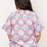 Woman with dark hair standing in a back profile position wearing the Mia Top against a white background. The focal point of this image is the intricate blue and pink floral design as well as pleating on collar and sleeves. The model paired this top with light-wash denim jeans.