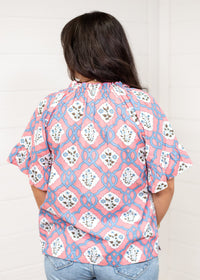 Woman with dark hair standing in a back profile position wearing the Mia Top against a white background. The focal point of this image is the intricate blue and pink floral design as well as pleating on collar and sleeves. The model paired this top with light-wash denim jeans.