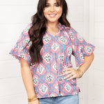 Woman with dark hair standing in a front profile position wearing the Mia Top against a white background. The focal point of this image is the intricate floral pink and blue pattern with tassels and puffy sleeves. The model paired this top with light-wash denim jeans. 