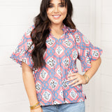Woman with dark hair standing in a front profile position wearing the Mia Top against a white background. The focal point of this image is the intricate floral pink and blue pattern with tassels and puffy sleeves. The model paired this top with light-wash denim jeans. 