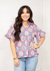 Woman with dark hair standing in a front profile position wearing the Mia Top against a white background. The focal point of this image is the intricate floral pink and blue pattern with tassels and puffy sleeves. The model paired this top with light-wash denim jeans. 