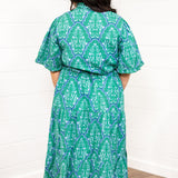 Woman with dark hair standing in a back profile position wearing the Nova Dress against a white background. The focal point of this image is the blue/green graphics on the dress. 