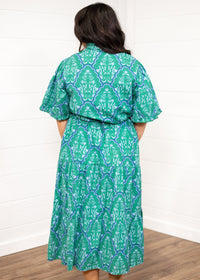 Woman with dark hair standing in a back profile position wearing the Nova Dress against a white background. The focal point of this image is the blue/green graphics on the dress. 