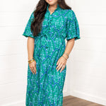 Woman with dark hair standing in a front profile position wearing the Nova Dress against a white background. The focal point of this image is the front details on dress (ruffle down buttons, belt). 