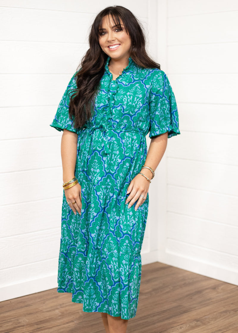 Woman with dark hair standing in a front profile position wearing the Nova Dress against a white background. The focal point of this image is the front details on dress (ruffle down buttons, belt). 