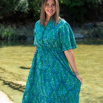 Woman with dark hair standing in a front profile position wearing the Nova Dress in a lifestyle scene. The focal point of this image is to see the floral patterns against greenery/water. 