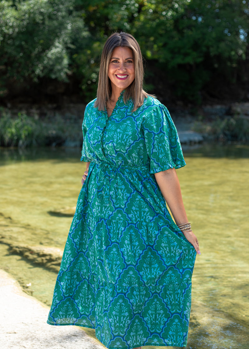 Woman with dark hair standing in a front profile position wearing the Nova Dress in a lifestyle scene. The focal point of this image is to see the floral patterns against greenery/water. 
