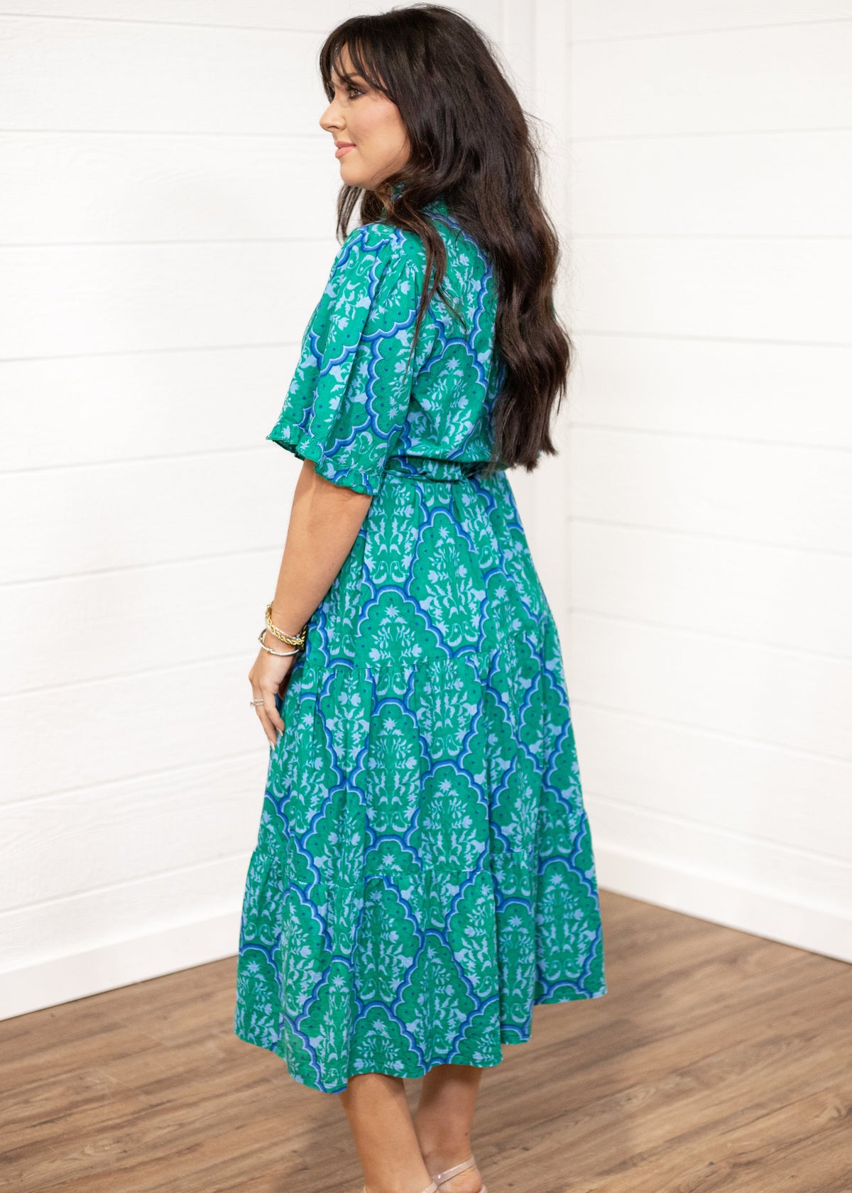 Woman with dark hair standing in a side profile position wearing the Nova Dress against a white background. The focal point of this image is the graphics on the dress as well as the sleeve length and belt. 