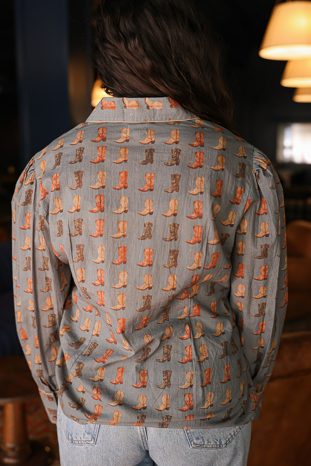 Woman with dark hair standing in a back profile position wearing the Oakley Top in a lifestyle scene. The focal point of this image is the boot graphics. The model paired this top with light-wash denim jeans. 