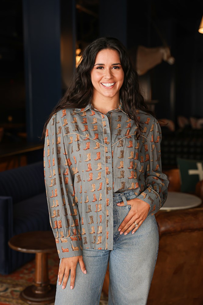 Woman with dark hair standing in a front profile position wearing the Oakley Top in a lifestyle scene. The focal point of this image is the boot graphics, buttons down front, and outlining on chest, collar, and cuff. The model paired this top with light-wash denim jeans. 