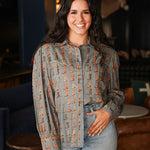 Woman with dark hair standing in a front profile position wearing the Oakley Top in a lifestyle scene. The focal point of this image is the boot graphics, buttons down front, and outlining on chest, collar, and cuff. The model paired this top with light-wash denim jeans. 