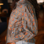 Woman with dark hair standing in a side profile position wearing the Oakley Top in a lifestyle scene. The focal point of this image is the boot graphics on sleeves and front of top, and the outlining on cuff, chest, and collar. The model paired this top with light-wash denim jeans.