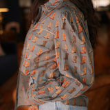 Woman with dark hair standing in a side profile position wearing the Oakley Top in a lifestyle scene. The focal point of this image is the boot graphics on sleeves and front of top, and the outlining on cuff, chest, and collar. The model paired this top with light-wash denim jeans.