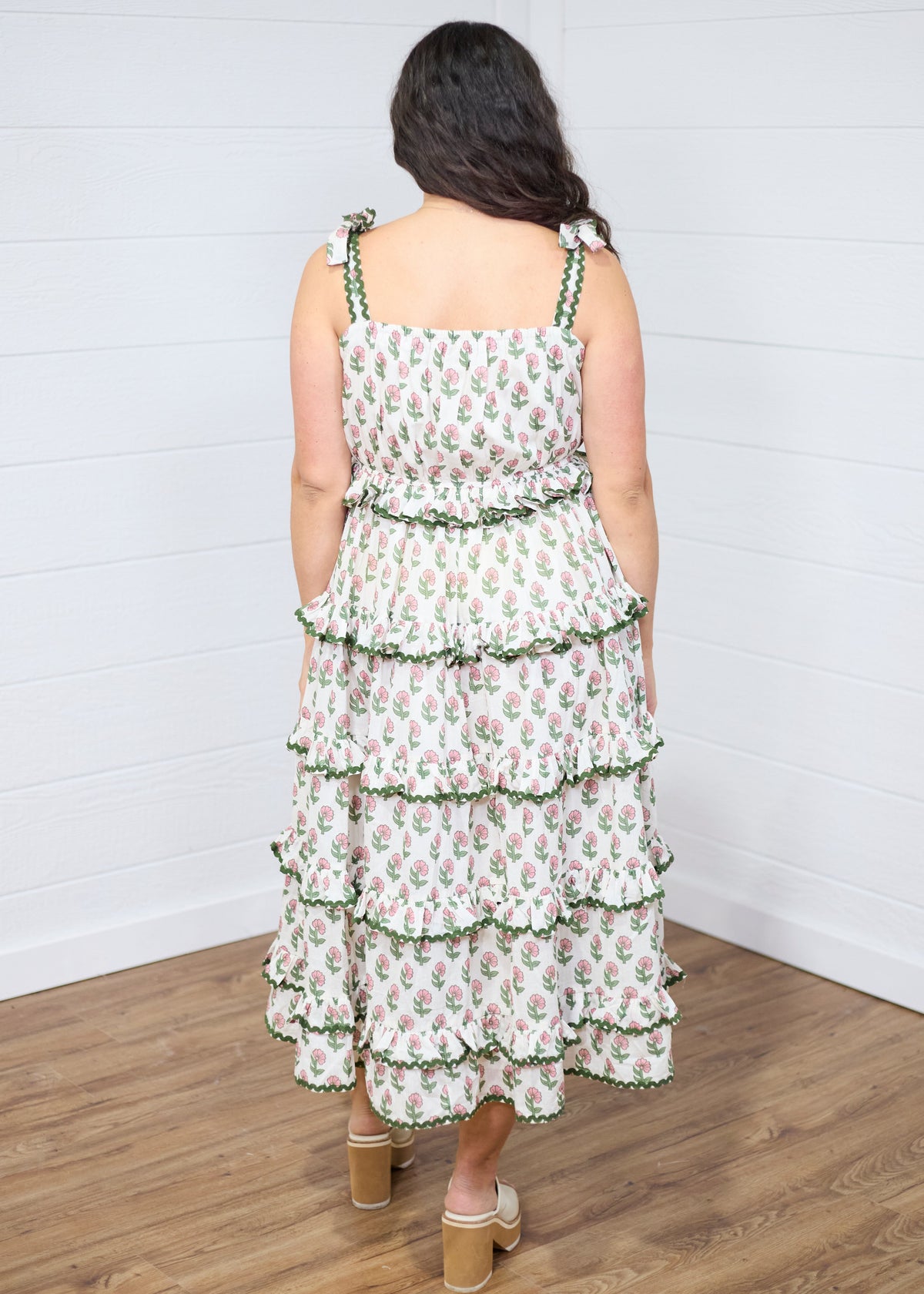 Woman with dark hair standing in a back profile position wearing the Paloma Dress against a white background. The focal point of this image is the floral graphics and tiered ruffles. The model paired this dress with cream heels. 