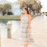 Woman with dark hair standing in a front profile position wearing the Paloma Dress in a lifestyle scene. The focal point of this image is to see the dress in its entirety including all graphics and length. The model paired this dress with white strappy heels. 