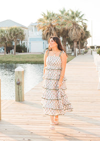 Woman with dark hair standing in a front profile position wearing the Paloma Dress in a lifestyle scene. The focal point of this image is to see the dress in its entirety including all graphics and length. The model paired this dress with white strappy heels. 