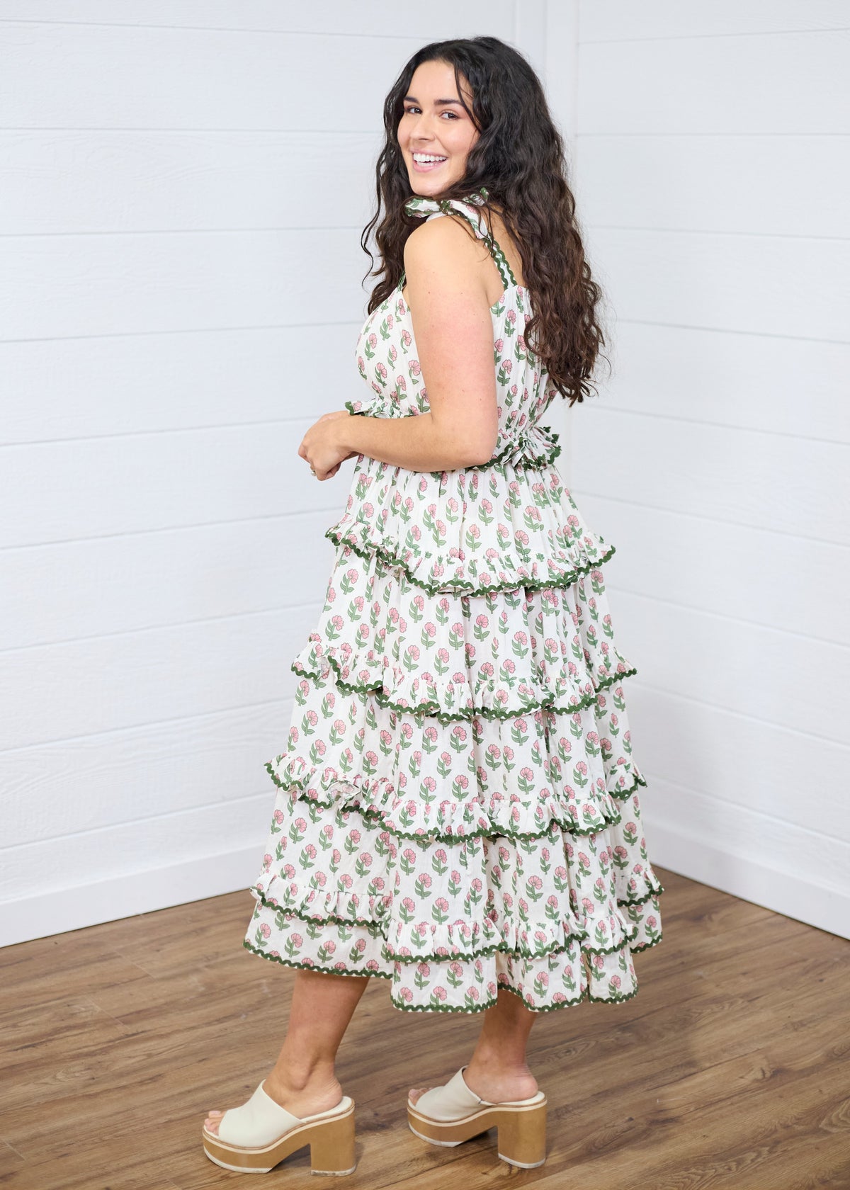 Woman with dark hair standing in side profile position wearing the Paloma Dress against a white background. The focal point of this image is the side ruffles of the dress, the length, and graphics. The model paired this dress with cream heels. 