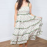 Woman with dark hair standing in a front profile position wearing the Paloma Dress against a white background. The focal point of this image is the ruffles, graphics, embroidered edges, and embroidered edges of the tied straps. The model paired this dress with cream heels. 