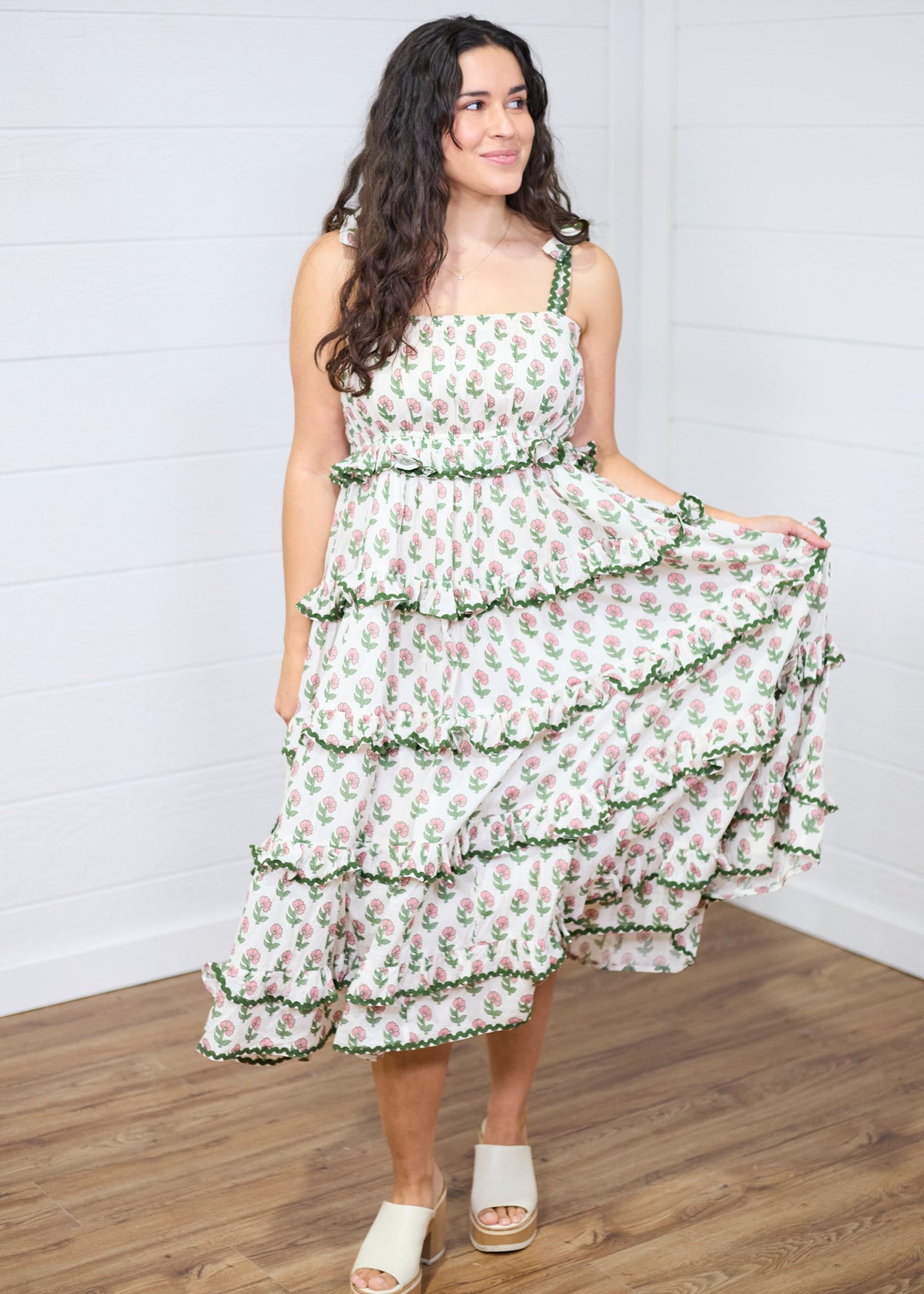 Woman with dark hair standing in a front profile position wearing the Paloma Dress against a white background. The focal point of this image is the ruffles, graphics, embroidered edges, and embroidered edges of the tied straps. The model paired this dress with cream heels. 
