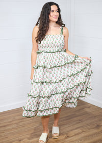 Woman with dark hair standing in a front profile position wearing the Paloma Dress against a white background. The focal point of this image is the ruffles, graphics, embroidered edges, and embroidered edges of the tied straps. The model paired this dress with cream heels. 