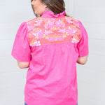Woman with dark hair standing in a back profile position wearing the Reece Top against a white background. The focal point of this image is the orange, yellow, and white embroidery on back of shoulder, pleating and ruffle collar. The model paired this top with dark-wash denim shorts. 