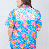 Woman with dark hair standing in a back profile wearing the Reece Top against a white background. The focal point of this image is the Hawaiian print as well as the white embroidery across the shoulders. The model paired this top with dark-wash denim shorts. 