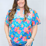 Woman with dark hair standing in front profile position wearing the Reece Top against a white background. The focal point of this image is the embroidery across chest, Hawaiian print, and the v-neckline. The model paired this top with dark-wash denim shorts.