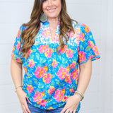 Woman with dark hair standing in front profile position wearing the Reece Top against a white background. The focal point of this image is the embroidery across chest, Hawaiian print, and the v-neckline. The model paired this top with dark-wash denim shorts.