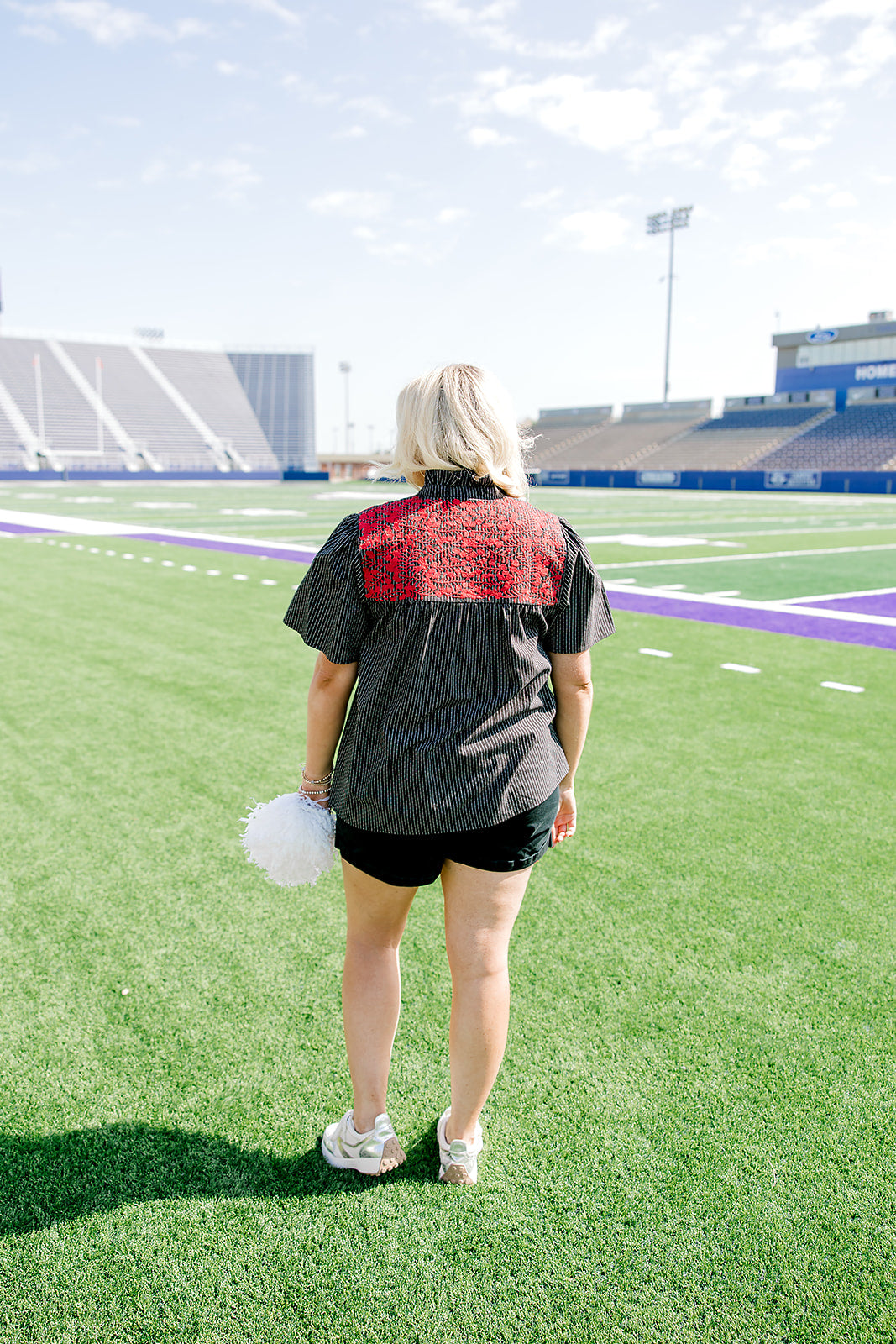 Woman with blonde hair standing in a back profile position wearing the Reece Top in a lifestyle scene. The focal point of this image is the red floral embroidery on back of top as well as pleating on back. The model paired this top with black denim shorts. 